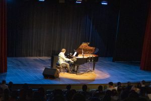 No palco do Teatro SARAH Brasília, de terno azul e sentado ao piano, maestro e pianista Eduardo Lages acompanha a cantora Marina Elali, de pé e de vestido longo verde, em apresentação de grandes sucessos de Roberto Carlos e Erasmo Carlos.