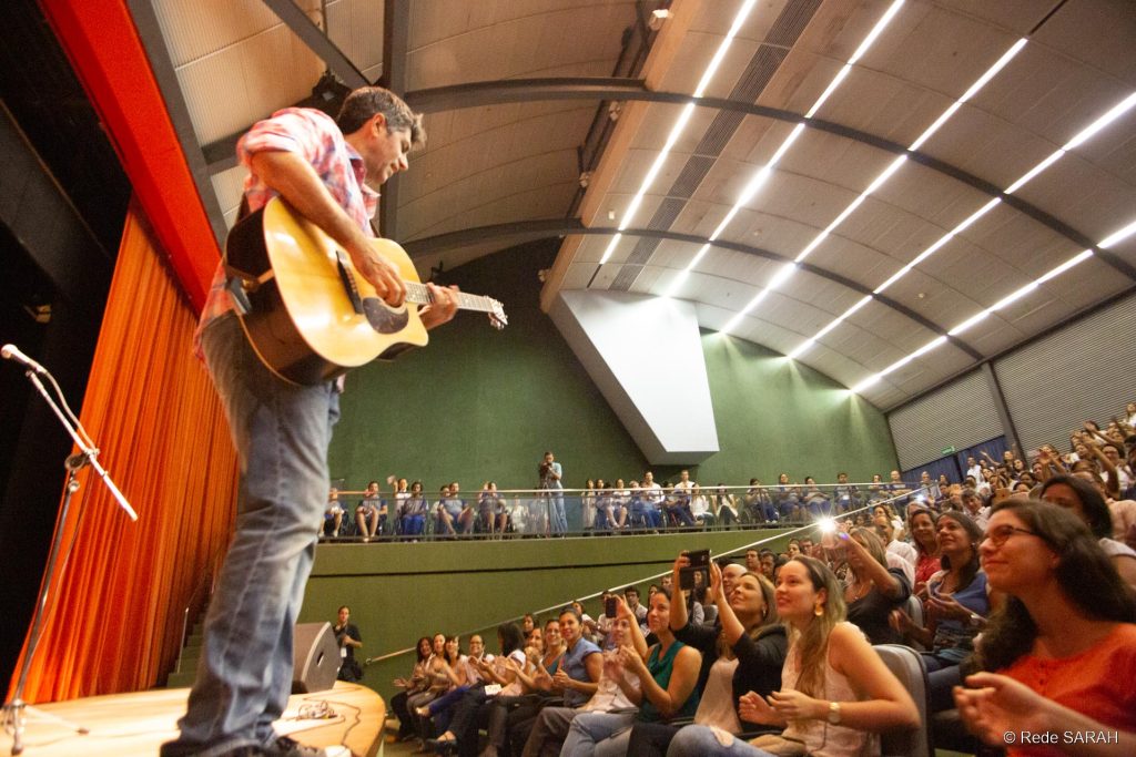 À esquerda, de pé no palco do Teatro SARAH, Leoni toca violão e canta. À direita e ao fundo, plateia de pacientes, acompanhantes e colaboradores da Rede SARAH bate palmas, canta e tira fotos com celulares.
