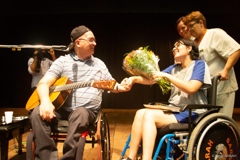 À esquerda, sentado em cadeira de rodas, no palco iluminado do Teatro SARAH, trajando camisa polo listrada azul, calça jeans preta e boné preto com a aba voltada para trás, e segurando um violão embaixo do braço direito, Herbert Vianna aperta a mão de jovem paciente, que lhe entrega um buquê de flores. Ela, trajando uniforme hospitalar e também sentada em cadeira de rodas, sorri para o artista. À direita e mais atrás, acompanhante e colaboradora, ambas trajando uniformes, acompanham a cena e sorriem.