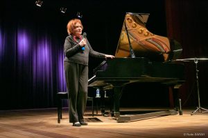 De pé, no palco do Teatro SARAH, trajando roupas pretas e segurando microfone com a mão direita, a pianista Clara Sverner fala à plateia.