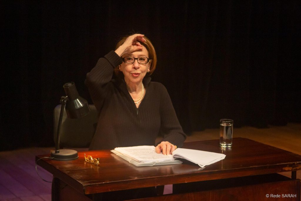 Sentada a uma mesa de madeira escura e trajando um pulôver preto, no centro do palco iluminado do Teatro SARAH, Fernanda Montenegro faz uma leitura dramática de textos de Nelson Rodrigues.