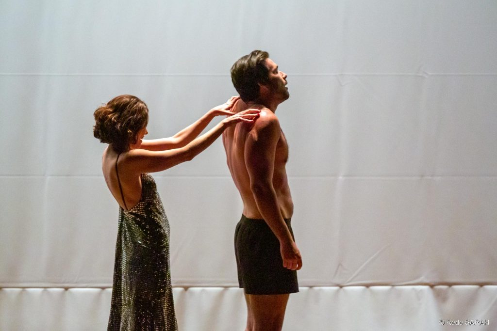 De pé, no palco do Teatro SARAH, em frente a um fundo branco, Juliana Martins, trajando um vestido longo de lantejoulas verdes, aparenta massagear as costas de Sérgio Marone, vestindo cueca tipo samba canção.