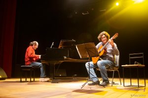 O pianista português João Lucas (esq.), trajando camisa social de mangas compridas vermelha e calças jeans, e o violonista brasileiro Jaime Ernest Dias, trajando camiseta de algodão de mangas longas cinza e calças jeans, apresentam-se no centro do palco iluminado do Teatro SARAH Brasília.