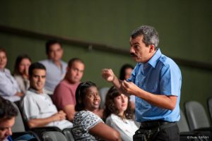 De pé, o ator Alexandre Lino, trajando uniforme de porteiro, com camisa de mangas curtas azul e calças pretas, aproxima-se da plateia de pacientes, acompanhantes e colaboradores da Rede enquanto atua no Teatro SARAH.