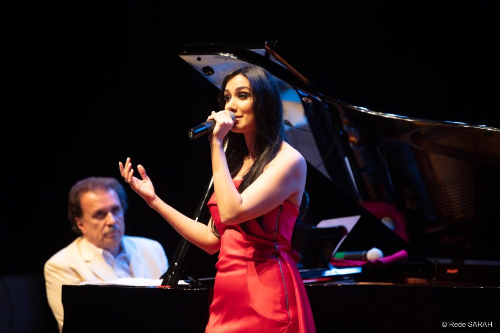 Trajando um vestido longo de cetim vermelho, Marina Elali canta ao microfone, no palco do Teatro SARAH. Ao fundo, o maestro Eduardo Lages acompanha ao piano cauda.