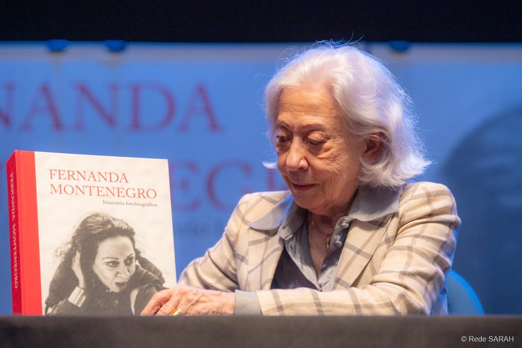Sentada a uma mesa, no palco do Teatro SARAH Brasília, Fernanda Montenegro segura uma cópia do seu livro "Itinerário Fotobiográfico".