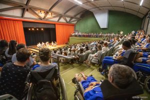 Em primeiro plano, nas galerias laterais e no auditório do Teatro SARAH, pacientes, acompanhantes e colaboradores da Rede, todos vestindo uniformes ou jalecos, acompanham a apresentação do Quinteto de Sopro e Percussão. Ao fundo, no centro do palco iluminado, os músicos tocam seus instrumentos.