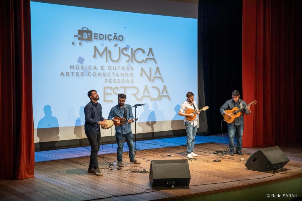 No palco iluminado do Teatro SARAH, de pé, apresentam-se quatro músicos, tocando pandeiro, cavaquinho, bandolim e violão. Trajam calças jeans, camisas de mangas compridas e tênis. Ao fundo, grande tela de projeção mostra os dizeres "8ª Edição – Música na Estrada, Música e Outras Artes Conectando as Pessoas”.