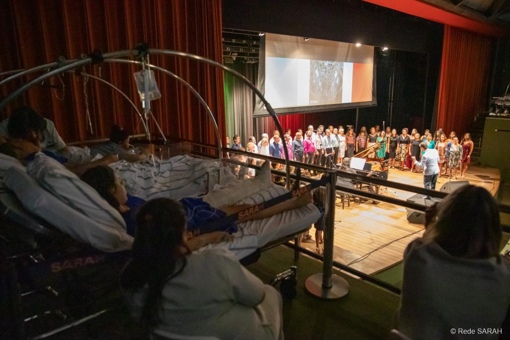 Em primeiro plano, em uma das galerias laterais do Teatro SARAH, à sombra, três pacientes, deitados em macas e trajando uniformes hospitalares, acompanham o concerto “ITÁLIA – Grazie Mille”. Ao seu lado, sentadas em cadeiras, duas acompanhantes também assistem ao espetáculo. Ao fundo, no centro do palco iluminado, apresenta-se o Coral TST-Capes, regido pelo maestro Carlos Ilha.