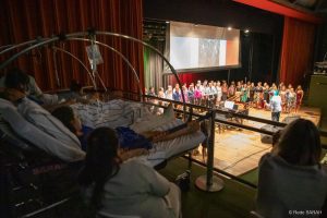 Em primeiro plano, em uma das galerias laterais do Teatro SARAH, à sombra, três pacientes, deitados em macas e trajando uniformes hospitalares, acompanham o concerto “ITÁLIA – Grazie Mille”. Ao seu lado, sentadas em cadeiras, duas acompanhantes também assistem ao espetáculo. Ao fundo, no centro do palco iluminado, apresenta-se o Coral TST-Capes, regido pelo maestro Carlos Ilha.