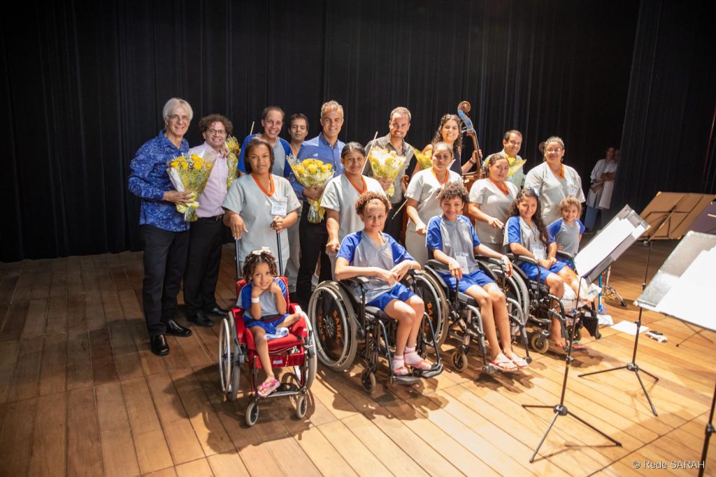 Na coxia do Teatro SARAH, organizados em fileiras, Ney Rosauro e o Quarteto Capital posam para fotos com pacientes e acompanhantes. Em primeiro plano, cinco pacientes cadeirantes, trajando uniformes hospitalares. Logo atrás, cinco acompanhantes, também vestindo uniformes hospitalares. Ao fundo, Ney Rosauro e os cinco músicos do Quarteto Capital seguram ramalhetes de flores.