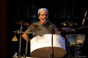 Close mostra o percussionista Sérgio Boré cantando ao microfone e tocando um tambor de aço. Iluminado, no palco do Teatro SARAH, ele traja camiseta cinza e boina azul-clara.