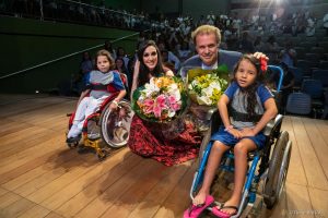 Logo após apresentação no Teatro SARAH, Eduardo Lages e Marina Elali, agachados e segurando ramalhetes de flores, posam com crianças pacientes do SARAH Brasília, ambas em cadeiras de rodas, no palco.