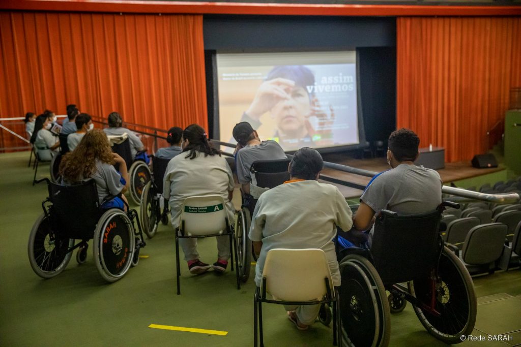 Em primeiro plano, em uma das galerias laterais do Teatro SARAH, pacientes em cadeiras de rodas, acompanhantes e colaboradores da Rede acompanham apresentação. Ao fundo, no palco, grande tela de projeção mostra imagem de uma mulher gesticulando em Libras e os dizeres "Assim Vivemos – 10º Festival Internacional de Filmes sobre Deficiência".