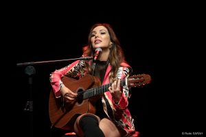 Sentada, Liah Soares canta e toca violão no Teatro SARAH. Ela traja um casado listrado, um vestido curto e botas pretas de cano longo e salto alto.