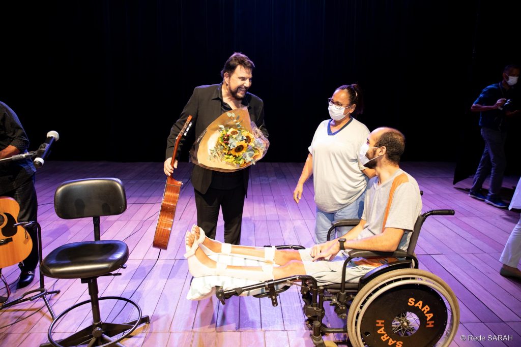 De pé, no palco do Teatro SARAH, trajando um terno e camisa social pretos, sem gravata, e segurando um violão na mão direita e um buquê de flores na esquerda, Michael Sullivan conversa com paciente em cadeira de rodas e sua acompanhante.