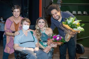 A atriz Maria Paula (à esquerda, vestindo calças jeans, uma bata estampada laranja e rosa, e uma blusa marrom), uma paciente do SARAH Brasília (sentada em cadeira de rodas, vestindo uniforme hospitalar), a cantora Juliani Fernandes (agachada, segurando um buquê de flores e trajando um vestido longo azul, verde e branco) e o violonista Diego Figueiredo (vestindo camisa de mangas longas azul e calças jeans) posam para foto no Teatro SARAH.