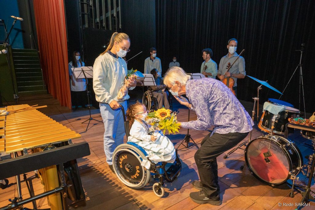 Na coxia do Teatro SARAH, vestindo calças pretas e camisa de mangas compridas roxa com estampa floral, Ney Rosauro abaixa-se para receber flores de criança em cadeira de rodas, paciente do SARAH Brasília. Mais atrás, acompanhante da criança observa a cena. Ao fundo aparecem membros do Quarteto Capital, segurando seus instrumentos.