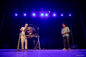 De pé e envolto por luzes azuis, no centro do palco do Teatro SARAH Brasília, o percussionista Sérgio Boré toca um tambor de aço, junto com outro percussionista, não identificado, que toca pandeiro, também de pé.
