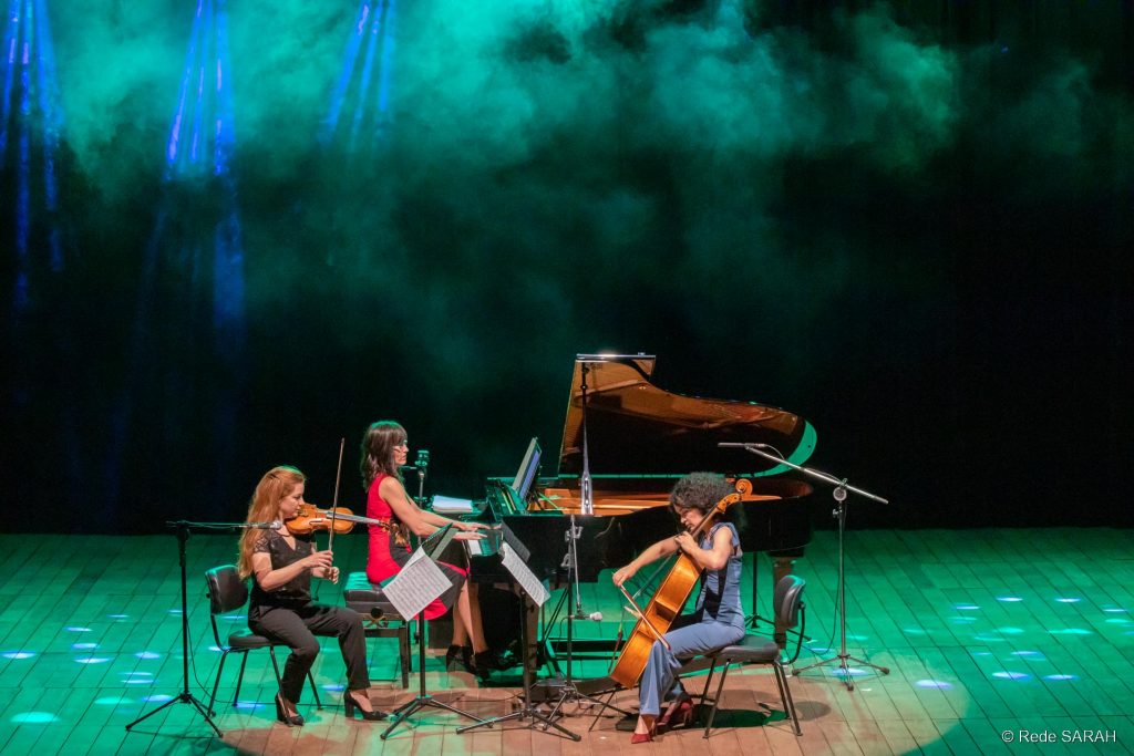 Envoltas em fumaça de gelo seco e luzes azuis e verdes, pianista, violinista e violoncelista apresentam-se no palco do Teatro SARAH em Brasília.