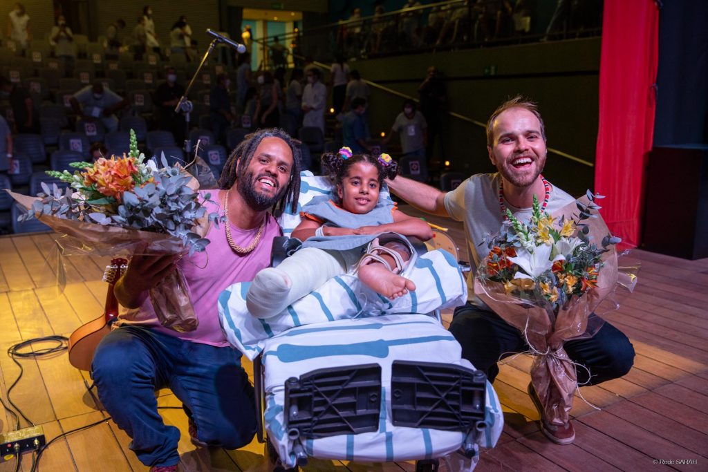 O bandolinista Filipe Braga e o violonista Stênio Neves, agachados e segurando buquês de flores, posam, no palco do Teatro SARAH, com paciente do SARAH Brasília, garota deitada em maca, com uniforme do hospital e uma das pernas engessada.