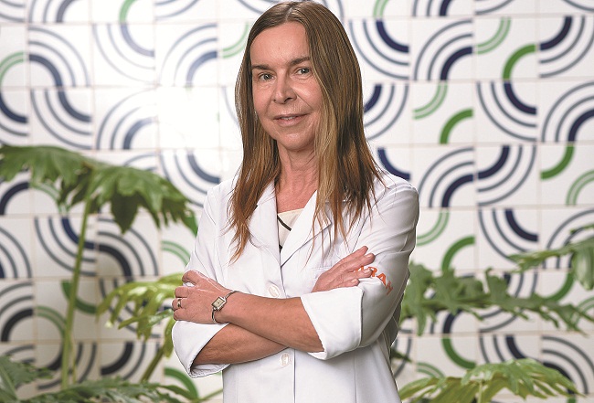 De pé e vestindo jaleco branco, Dra. Lúcia Willadino Braga posa para foto, de braços cruzados, em frente a azulejos de Athos Bulcão.