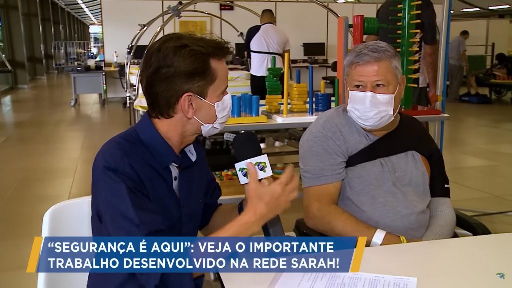 Sentado, à esquerda, em frente a uma mesa e vestindo camisa azul-marinho e máscara cirúrgica, jornalista entrevista paciente, sentado em cadeira de rodas, vestindo uniforme da Rede SARAH e máscara cirúrgica. Ao fundo, pacientes e profissionais da saúde realizam exercícios de reabilitação.