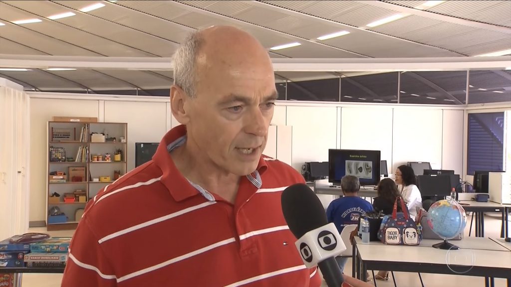 De pé e vestindo camisa polo vermelha, com listras brancas, homem fala a repórter da TV Globo no SARAH Lago Norte, em Brasília. Ao fundo, pacientes a profissionais de reabilitação conversam entre si.