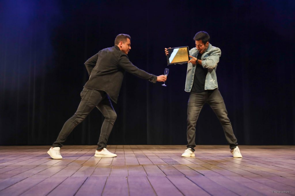 Ilusionistas Henry & Klauss apresentam-se no Teatro SARAH. Um deles segura uma taça vazia de espumante, enquanto o outro segura a tela de um tablet e parece derramar o conteúdo da tela na taça.