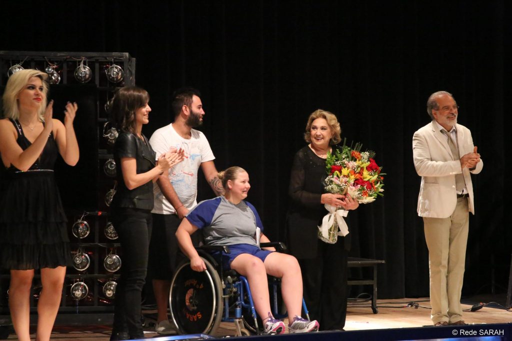De pé e segurando um grande buquê de flores que acabou de receber de uma paciente sentada em cadeira de rodas, no palco do Teatro SARAH, Eva Wilma recebe palmas dos demais membros do elenco.