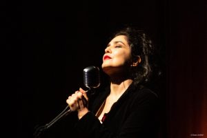 Close mostra Letícia Sabatella, iluminada por um spot de luz, cercada de sombras, vestindo um sobretudo preto, cantando, de olhos fechados, enquanto segura microfone e pedestal.