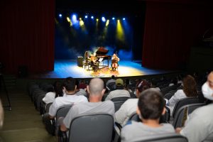 Em primeiro plano, sentados nas poltronas do Teatro SARAH e de costas, aparecem pacientes, acompanhantes e colaboradores da Rede SARAH. Ao fundo, no centro do palco iluminado, apresentam-se pianista, violinista e violoncelista.