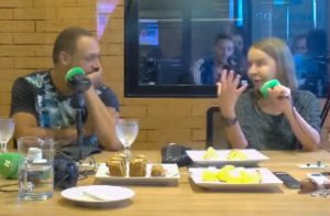 Captura de tela mostra imagem da transmissão, ao vivo, do programa de rádio Mesa pra Dois: sentada, à direita, Dra. Lúcia Willadino Braga conversa com membros da banda Moinho d’Água, todos sentados, em frente a microfones, ao redor de uma mesa com bebidas e lanche.