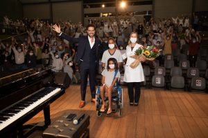 Em fotografia tirada do fundo do palco do Teatro SARAH, o tenor Thiago Arancam, vestindo terno azul-marinho escuro e camisa social branca, posa com a presidente da Rede SARAH, vestindo jaleco branco, e uma paciente, em cadeira de rodas, e sua acompanhante. Ao fundo, pacientes, acompanhantes e colaboradores da Rede acenam da plateia para a foto.