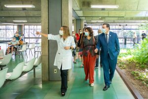 Vestindo jaleco branco, Dra. Lúcia Willadino Braga apresenta o ambulatório do SARAH Brasília Centro à ministra da Saúde, Nísia Trindade, vestindo blusa de manga comprida em tons de vermelho e azul com calças vermelhas, e ao secretário-executivo da Saúde, Swedenberger Barbosa, vestindo terno azul e gravata. Ao fundo, membros da comitiva ministerial e da equipe do hospital acompanham a visita.