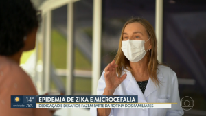 De pé, trajando jaleco branco e usando máscara cirúrgica, a presidente da Rede SARAH, Dra. Lúcia Willadino Braga, dá entrevista a repórter, que aparece de costas e desfocada.
