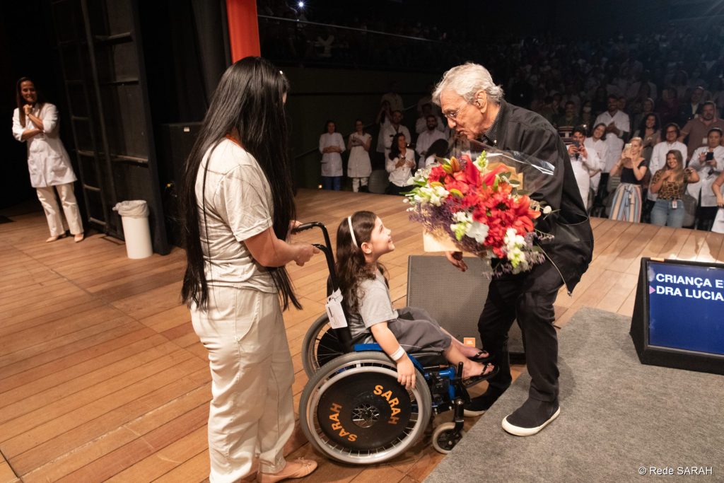 De pé e sorrindo, Caetano Veloso inclina-se para abraçar criança em cadeira de rodas, paciente do SARAH Brasília, de quem recebeu um buquê de flores.